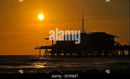 Pier Silhouette at Golden Ocean Sunset. Dark pier silhouette rises above rolling waves as the setting sun paints the ocean and sky in warm golden Banque D'Images