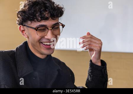 Homme adulte souriant, levant la main à l'intérieur, portant des verres de tortue, manteau, col roulé noir, boucle d'oreille Banque D'Images