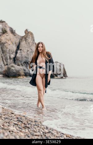 Femme plage océan marche le long de la rive rocheuse portant une élégante robe noire et maillot de bain beige pendant une journée brumeuse Banque D'Images