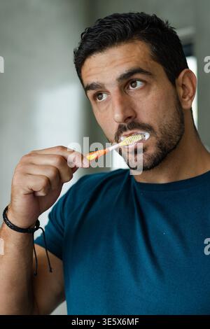 Homme en T-shirt se brossant les dents dans la salle de bain, tenant une brosse à dents orange-jaune avec mousse et bracelet Banque D'Images