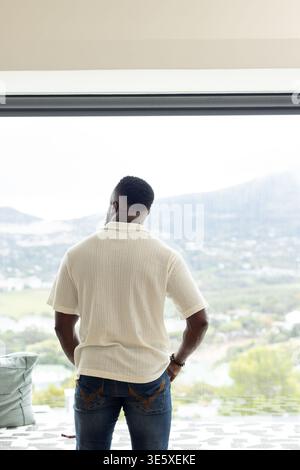Homme afro-américain debout par fenêtre dans une chambre moderne portant polo et jeans smartphone à proximité Banque D'Images