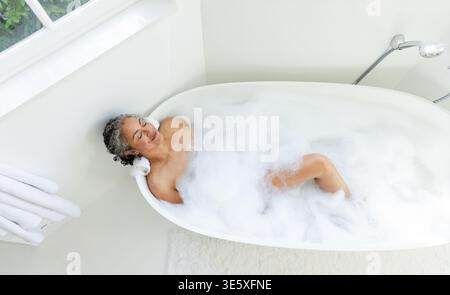 Femme afro-américaine s'inclinant dans la baignoire de la salle de bains, reposant la tête sur une serviette roulée avec de la mousse à bulles Banque D'Images