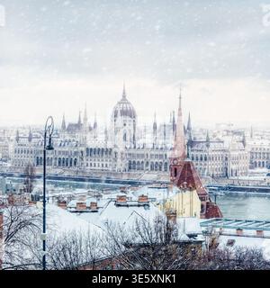 Bâtiment du Parlement hongrois vu à travers la neige tombant au-dessus des toits d'hiver et de la flèche de l'église sur le côté Buda de Budapest, Hongrie. Banque D'Images