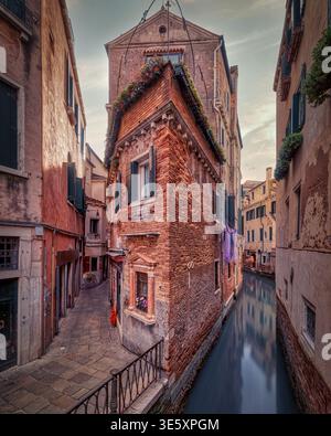 Canal vénitien étroit à côté de maisons en briques altérées et une ruelle tranquille dans la lumière chaude du soir, Venise, Italie. Banque D'Images