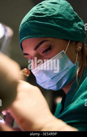 Chirurgien féminin focalisé portant un masque chirurgical et une casquette effectuant une procédure délicate en salle d'opération Banque D'Images
