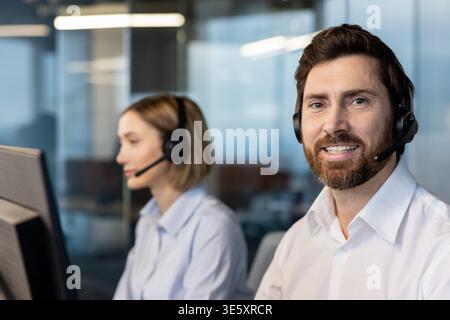 Agent de centre d'appels souriant, barbe, portant un casque et regardant la caméra tout en travaillant à côté d'une collègue sur un ordinateur dans un bureau moderne aidant les clients Banque D'Images