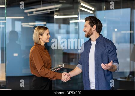 Deux professionnels du commerce souriants, une femme tenant une tablette et un homme portant une chemise en denim, serrant la main dans un bureau en verre moderne, symbolisant l’accord, le succès et la confiance dans leur nouvelle collaboration Banque D'Images