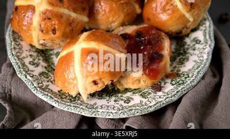 Gros plan d'un pain croisé glacé coupé en deux avec de la confiture de fruits noirs et des raisins secs. Les petits pains ont une texture dorée douce et sont servis sur un g. Banque D'Images