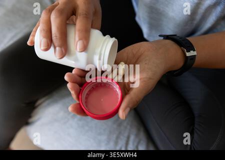 Vidéo verticale : dévisser la bouteille de pilule, femme versant des capsules dans la paume sur le canapé, portant le dessus gris Banque D'Images