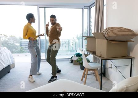 Danse couple diversifié filant dans la chambre avec portes de balcon, avec lit, boîtes de bureau et plante Banque D'Images