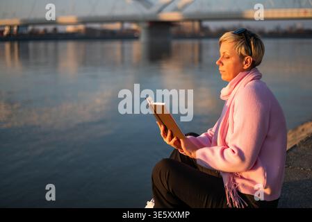 Femme blonde avec des cheveux courts lisant un livre tout en profitant assis confortablement par la rivière dans la ville. Banque D'Images