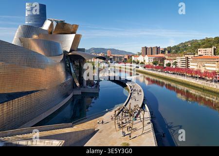 Les visiteurs se promènent le long de la rive de Bilbao près du musée Guggenheim. Les bâtiments reflètent un design moderne sous un ciel bleu clair. Banque D'Images