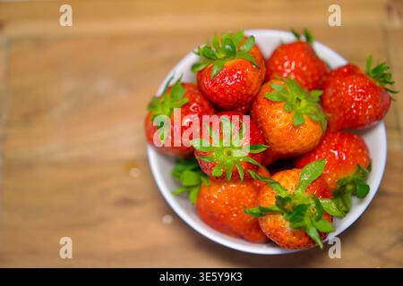 Vue en grand angle d'un bol en céramique blanche rempli à ras bord de grandes fraises mûres aux tiges vertes. Le bol repose sur un comptoir en bois naturel. La lumière du jour douce souligne la couleur rouge éclatante des baies et leur texture naturelle. fraises fraîches dans un bol blanc sur une surface en bois, concept alimentaire biologique. Photo de haute qualité Banque D'Images