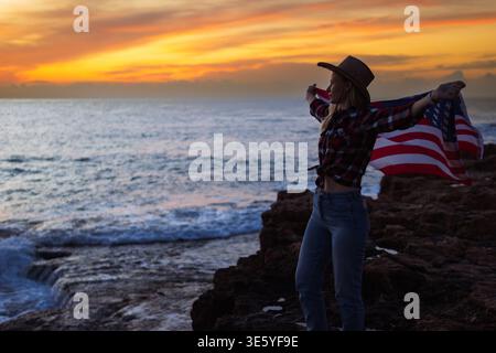 Femme portant chapeau de cow-boy et chemise à carreaux debout sur les rochers de l'océan avec drapeau américain soufflant dans le vent pendant le coucher du soleil, célébrant la liberté et Banque D'Images