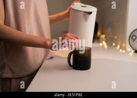 Gros plan de la jeune femme versant de l'eau chaude de la bouilloire électrique dans la tasse dans la cuisine. copier l'espace Banque D'Images