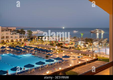 Hurghada, Égypte, 16 mars 2026 : un complexe de luxe éclairé au crépuscule avec une grande piscine, des parasols et des yachts ancrés dans le rouge Banque D'Images
