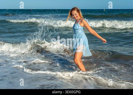 Jeune femme dans une robe bleu clair se sentant rafraîchie par éclaboussures de vagues de l'océan sur le rivage, profitant d'une journée de détente et de plaisir pendant son été Banque D'Images