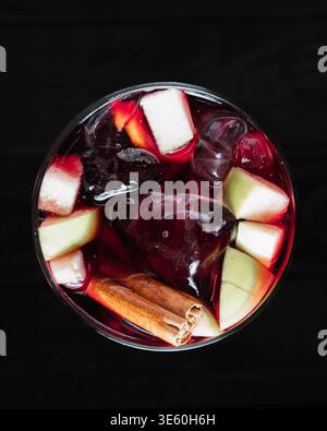 Le verre foncé contient du vin rouge, de la glace, des fruits, de la cannelle. Les bâtonnets de cannelle, les morceaux de pomme et les cubes flottent. Un liquide rouge foncé brille sous les fruits. Dos noir Banque D'Images