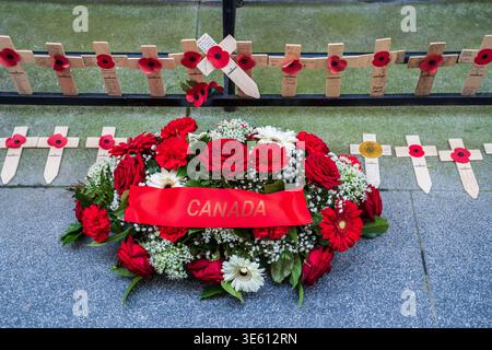 Une couronne de roses rouges et de gerberas avec un ruban 'CANADA' repose sur le sol. En arrière-plan, de petites croix en bois avec des coquelicots rouges sont alignées al Banque D'Images