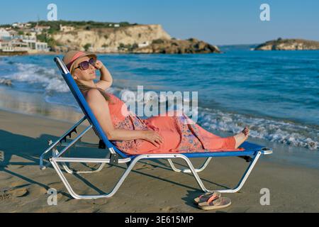 Une femme détendue dans une robe rose et un chapeau de soleil reposant sur une chaise longue bleue sur une plage de sable. Belles vagues de mer et côte rocheuse en arrière-plan. Banque D'Images