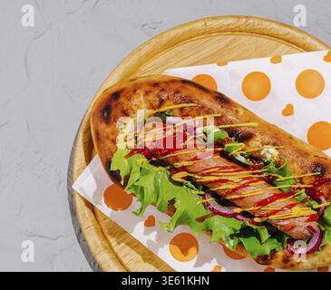 Nourriture, Un hot dog délicieux est présenté sur un plateau en bois, surmonté de laitue fraîche, tomates, cornichons et moutarde. Les couleurs vibrantes et la texture Banque D'Images