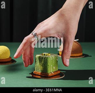 Nourriture, Un coup de main est atteint pour un dessert vert parmi diverses pâtisseries sur une table verte vibrante. Le cadre suggère un événement culinaire ou une dégustation exper Banque D'Images