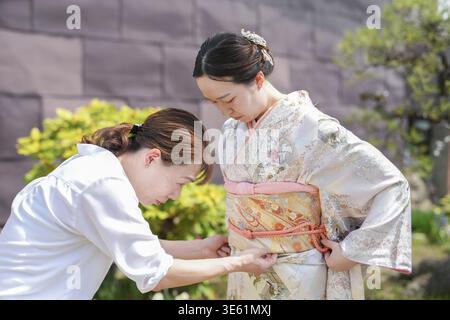 Une femme en kimono est aidée par une autre femme. La femme dans le kimono porte une tenue blanche et a ses bras autour du wais de l'autre femme Banque D'Images