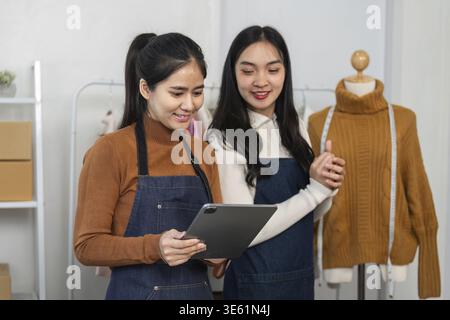Deux créatrices de mode qualifiées regardant une tablette, discutant de leurs dernières créations tout en se tenant debout à côté d'un présentoir de vêtements sur mesure dans un combat Banque D'Images