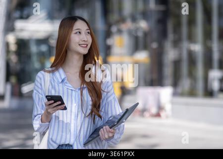Une femme marche dans la rue tenant un téléphone portable et un ordinateur portable. Elle sourit. L'ordinateur portable est dans sa main et le téléphone portable dans sa poche Banque D'Images