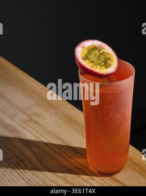 Nourriture, Un grand verre rempli d'un cocktail orange vibrant se trouve sur un plateau de bar en bois. Un fruit de la passion coupé en deux repose sur le bord, ajoutant une touche lumineuse à t Banque D'Images