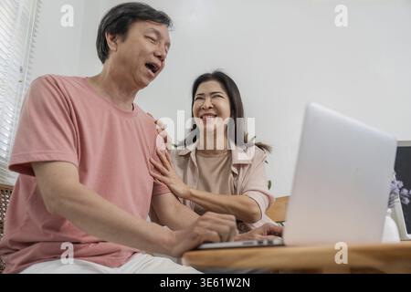 Un couple de personnes âgées joyeux profitant d'un moment léger tout en s'engageant avec un ordinateur portable, favorisant la connexion et le bonheur dans un cadre confortable à la maison Banque D'Images