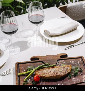Nourriture, steak grillé est présenté sur une planche de bois accompagné d'asperges et de tomates cerises. Deux verres de vin rouge sont assis sur une table magnifiquement dressée Banque D'Images