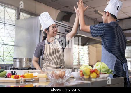 Deux jeunes chefs dans une cuisine partagent un joyeux high Five, célébrant le travail d'équipe et le succès dans la cuisine. L'environnement est chaleureux et rempli de frais Banque D'Images