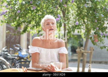 Une femme est assise à une table avec un verre d'eau devant elle. Elle porte une chemise blanche et a une expression sérieuse sur son visage. La scène est réglée Banque D'Images