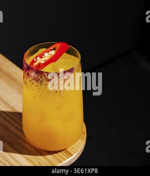 Nourriture, Un cocktail jaune vif se trouve sur une table en bois, garnie d'une tranche de piment. La boisson semble rafraîchissante et épicée contre un BA foncé Banque D'Images