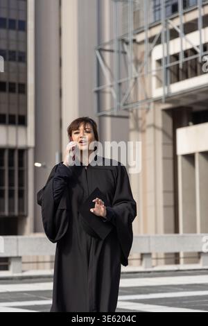 Femme dans la vingtaine en robe académique debout sur la plaza tenant le chapeau, parlant sur smartphone Banque D'Images