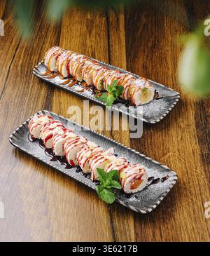 Nourriture, deux desserts exquis disposés sur des plats noirs, garnis de feuilles de menthe, servis sur une table en bois. Les rouleaux sont décorés avec du sirop A. Banque D'Images