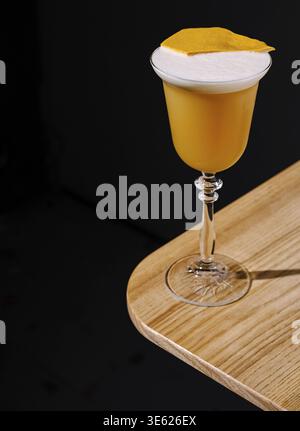 Food, Un cocktail jaune éclatant garni d'une tranche d'orange se trouve sur une table en bois élégante. La boisson présente un dessus mousseux, soulignant son refre Banque D'Images