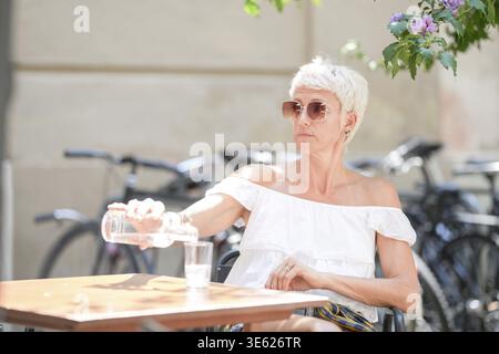 Une femme assise à une table avec un verre d'eau et une bouteille d'eau. Elle porte des lunettes de soleil et une chemise blanche Banque D'Images