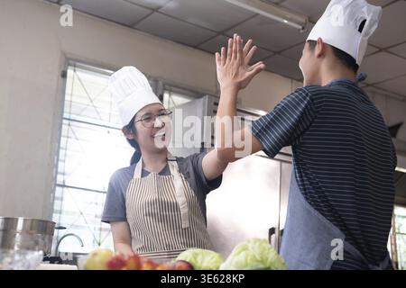 Deux jeunes chefs dans une cuisine partagent un joyeux high Five, célébrant le travail d'équipe et le succès dans la cuisine. L'environnement est chaleureux et rempli de frais Banque D'Images