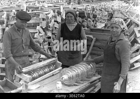 Slavyansk, années 1970, usine d'armature-isolant. Une équipe de trois travailleurs, un homme et deux femmes, emballe soigneusement de grands isolateurs de suspension en verre transparent dans des caisses en bois personnalisées. Le département d'emballage opère à l'extérieur, entouré de piles massives de cadres en bois jetés et de produits électriques industriels finis. Une authentique photographie d'archives historiques véhiculant l'esprit du travail de fabrication soviétique physique, la routine industrielle quotidienne authentique, la dynamique d'équipe et le Donbass paisible. Banque D'Images