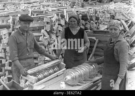 Slavyansk, années 1970, usine d'armature-isolant. Une équipe de trois travailleurs, un homme et deux femmes, emballe soigneusement de grands isolateurs de suspension en verre transparent dans des caisses en bois personnalisées. Le département d'emballage opère à l'extérieur, entouré de piles massives de cadres en bois jetés et de produits électriques industriels finis. Une authentique photographie d'archives historiques véhiculant l'esprit du travail de fabrication soviétique physique, la routine industrielle quotidienne authentique, la dynamique d'équipe et le Donbass paisible. Banque D'Images