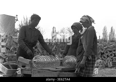 Slavyansk, années 1970, usine d'armature-isolant. Une équipe de trois travailleurs, un homme et deux femmes, sont engagés dans l'emballage manuel de grands isolateurs de suspension en verre transparent dans des caisses en bois. La zone extérieure de l'entrepôt est remplie de piles d'équipements électriques finis et de matériaux d'emballage en bois. Une authentique photographie d'archives historiques transmettant l'esprit du travail industriel soviétique physique collectif, la vie quotidienne de fabrication de routine, la dynamique d'équipe amicale et l'atmosphère du Donbass paisible. Banque D'Images
