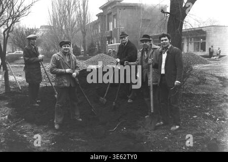 Slavyansk, 17 avril 1982, armature-isolateur usine. Une équipe de cinq ouvriers d'usine posent solennellement avec des pelles tout en se tenant autour d'un grand tas de terre sombre pendant le subbotnik communiste. Les hommes sont regroupés à l'extérieur dans la zone verte près du bâtiment de l'entreprise un jour de printemps. Une authentique photographie d'archives historiques véhiculant l'esprit confiant du travail bénévole collectif, la vie quotidienne industrielle soviétique authentique, la camaraderie professionnelle sur le lieu de travail et le Donbass paisible. Banque D'Images
