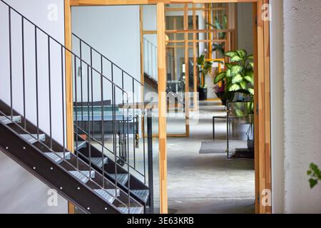 Intérieur de bureau moderne avec escalier en métal avec garde-corps verticaux au premier plan, vu à travers une cloison en verre encadrée de bois. Le Banque D'Images