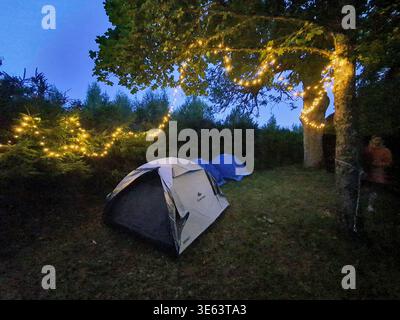 Une scène de camping au crépuscule avec deux tentes installées sur une zone herbeuse entourée d'arbres ornés de guirlandes lumineuses chaudes. Au premier plan, un blanc Banque D'Images