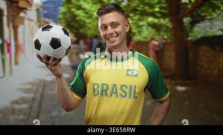 Man in brasil maillot tient le ballon de football avec la main droite et pointe le doigt tout en souriant dans la rue ; fierté joyeuse. Banque D'Images