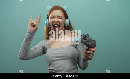 Femme rousse portant des écouteurs d'oreille de chat et tenant une manette de jeu, clignotants cornes de roche avec sa main et la langue en studio ; ludique. Banque D'Images