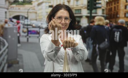 Femme hispanique d'âge moyen médecin en manteau blanc avec stéthoscope tient les poings en avant et sourit dans une rue animée ; résilience de confiance. Banque D'Images