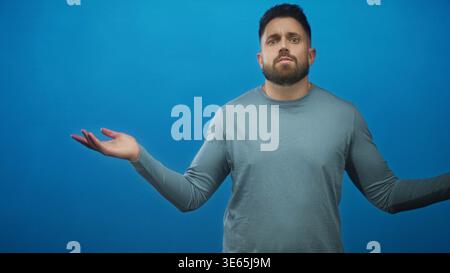 Homme à la barbe et aux mains visibles haussant les paumes vers le haut et levant les sourcils sur un fond de studio bleu sans couture ; incertitude. Banque D'Images